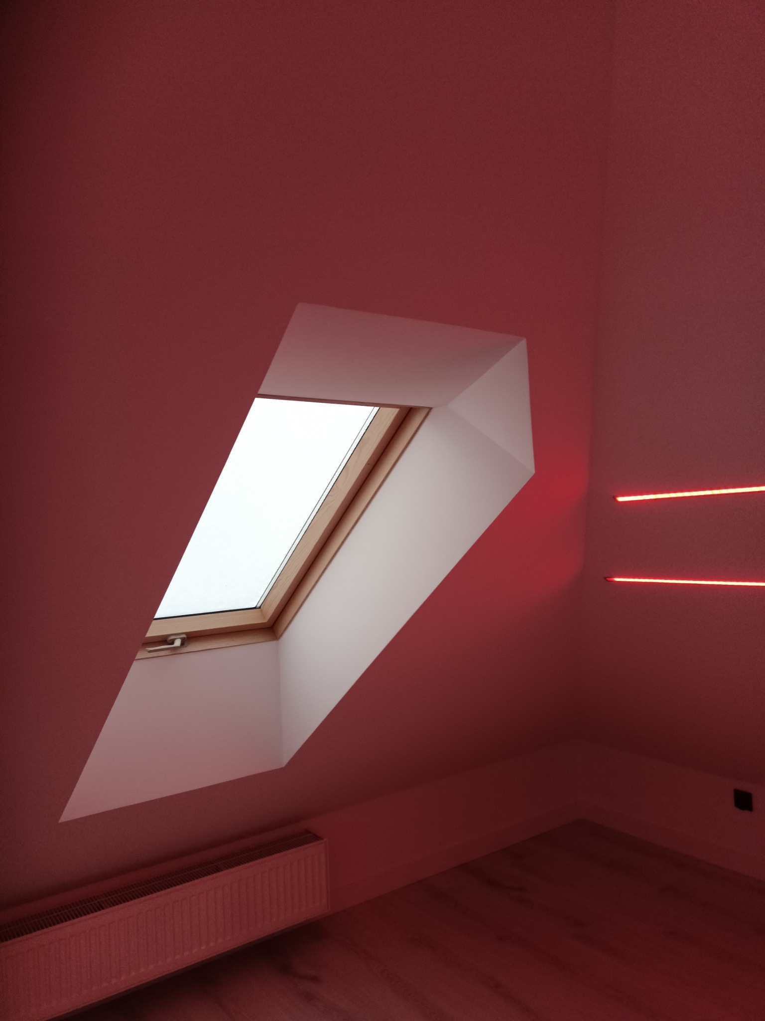 Poddasze po odnowieniu: skośne okno dachowe z drewnianą ramą, biały grzejnik, podłoga z paneli i czerwone oświetlenie LED na ścianie. Całość w ciepłej, czerwonej tonacji.