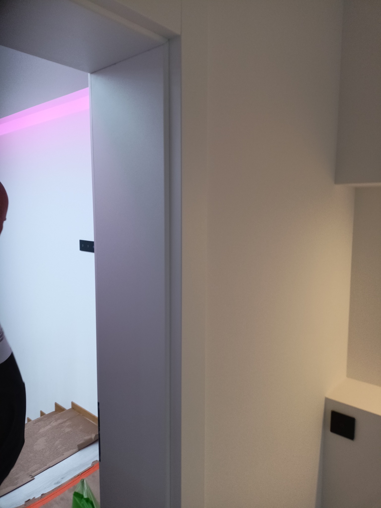 Nowoczesne wnętrze po remoncie: białe ściany, minimalistyczne drzwi z czarnymi zawiasami, widoczny fragment schodów i oświetlenie LED w kolorze różowym.