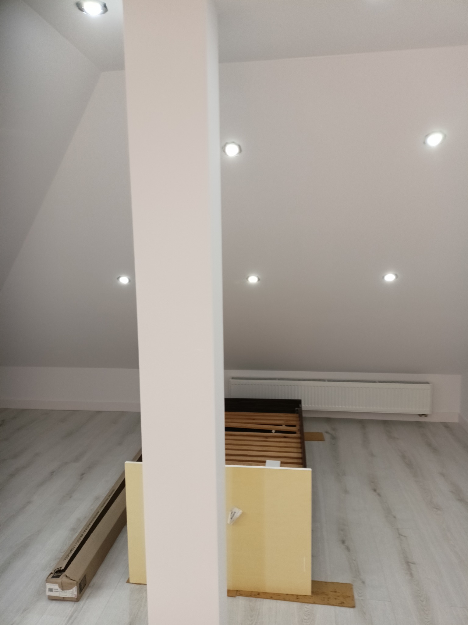 Poddasze z białymi ścianami i panelami, oświetlone punktowo. Widoczny fragment łóżka i karton, sugerujące etap prac wykończeniowych. Minimalistyczny styl.