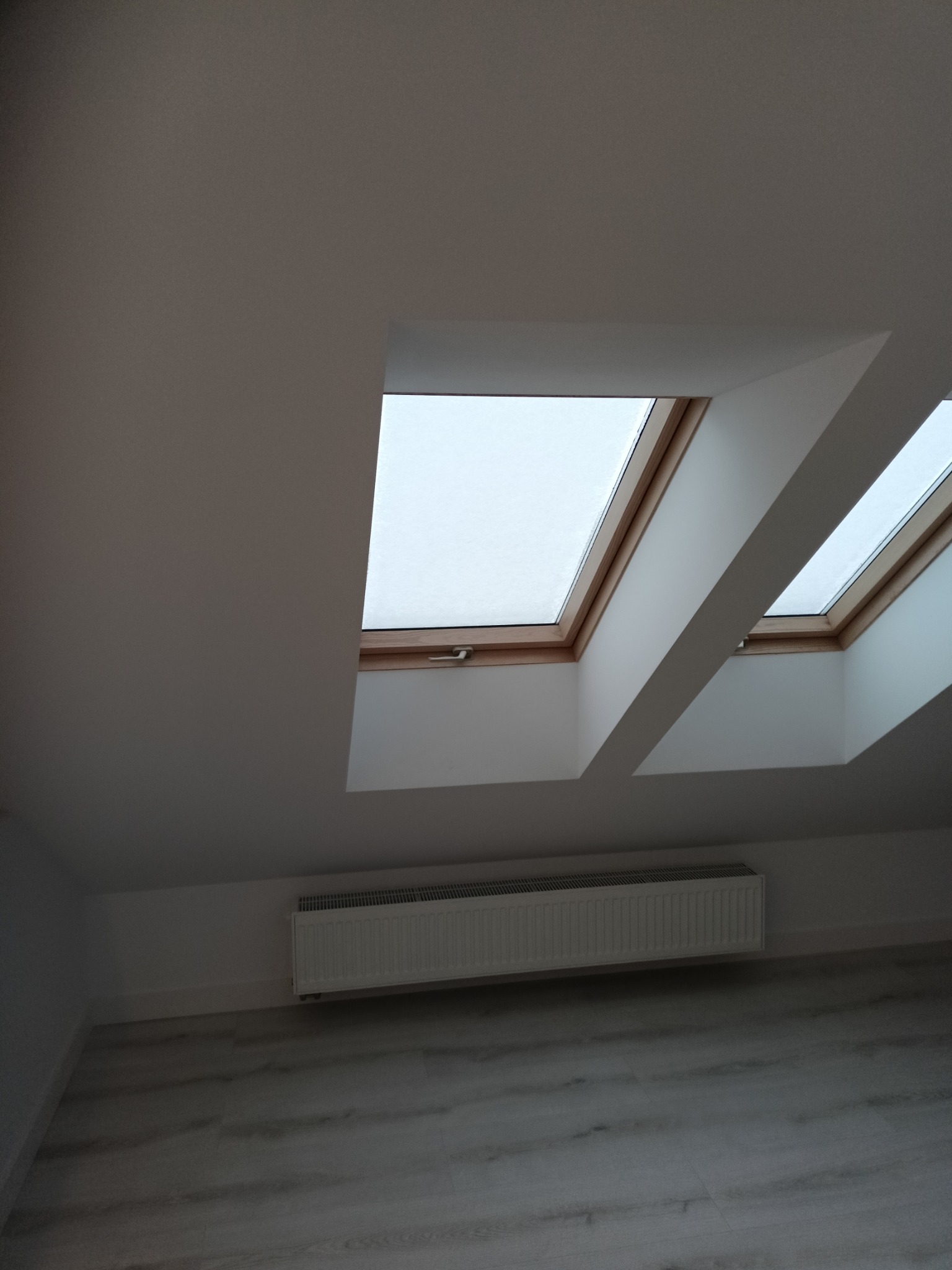 Jasne poddasze po remoncie z dwoma oknami dachowymi i grzejnikiem. Ściany i sufit pomalowane na biało, podłoga wyłożona panelami w odcieniach szarości.