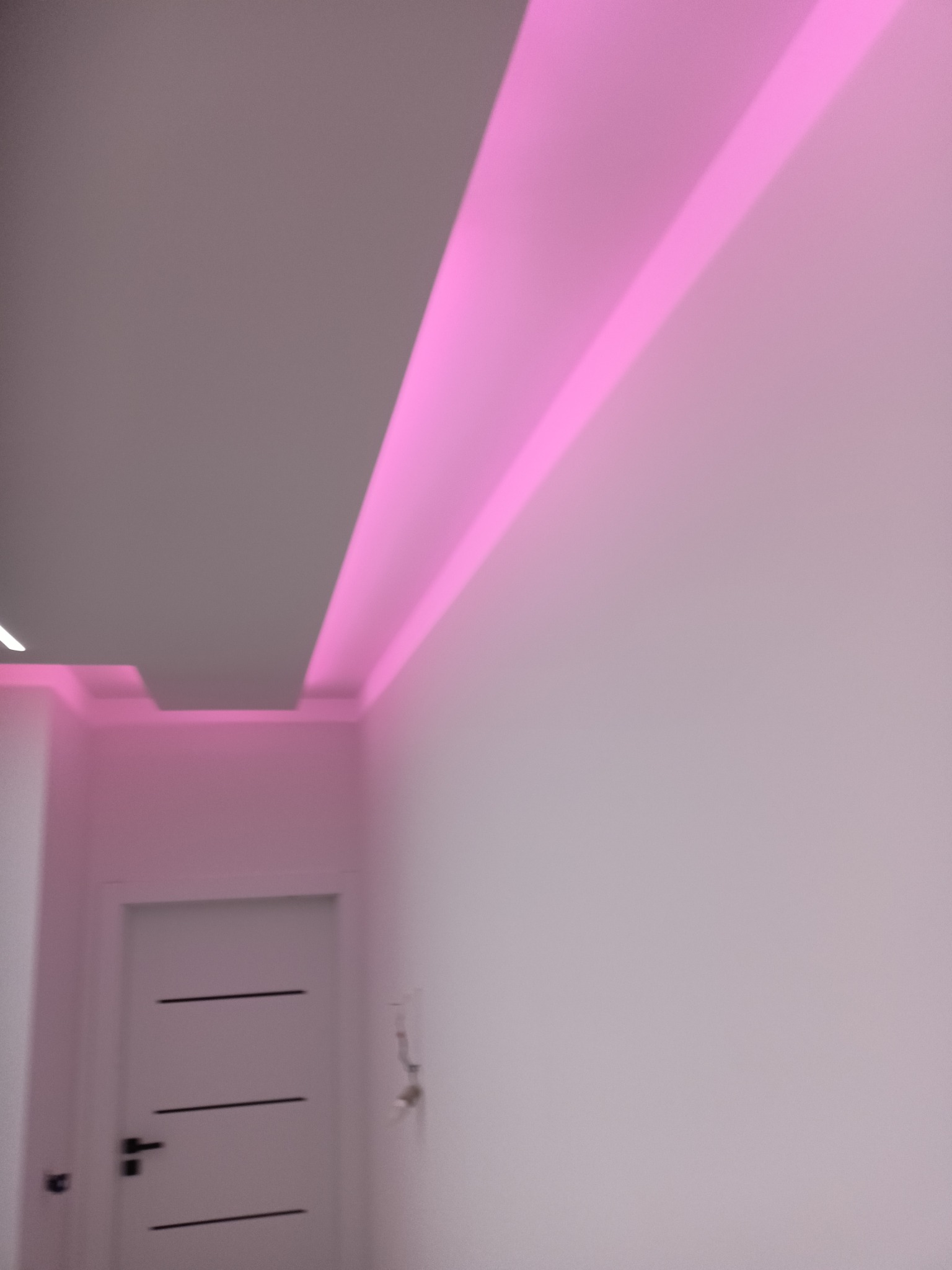 Białe ściany i sufit z podświetleniem LED w kolorze różowym, widoczny fragment drzwi. Nowoczesne wykończenie wnętrza z akcentem na oświetlenie.