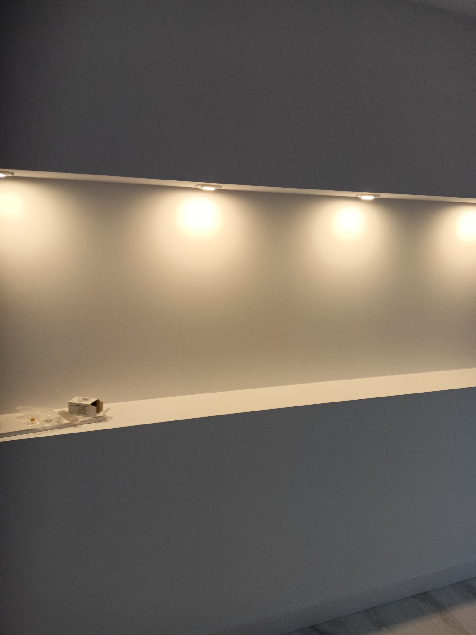 Ściana pomalowana na szaro z wnęką i oświetleniem punktowym. Na półce leżą akcesoria montażowe. Minimalistyczny design wnętrza.