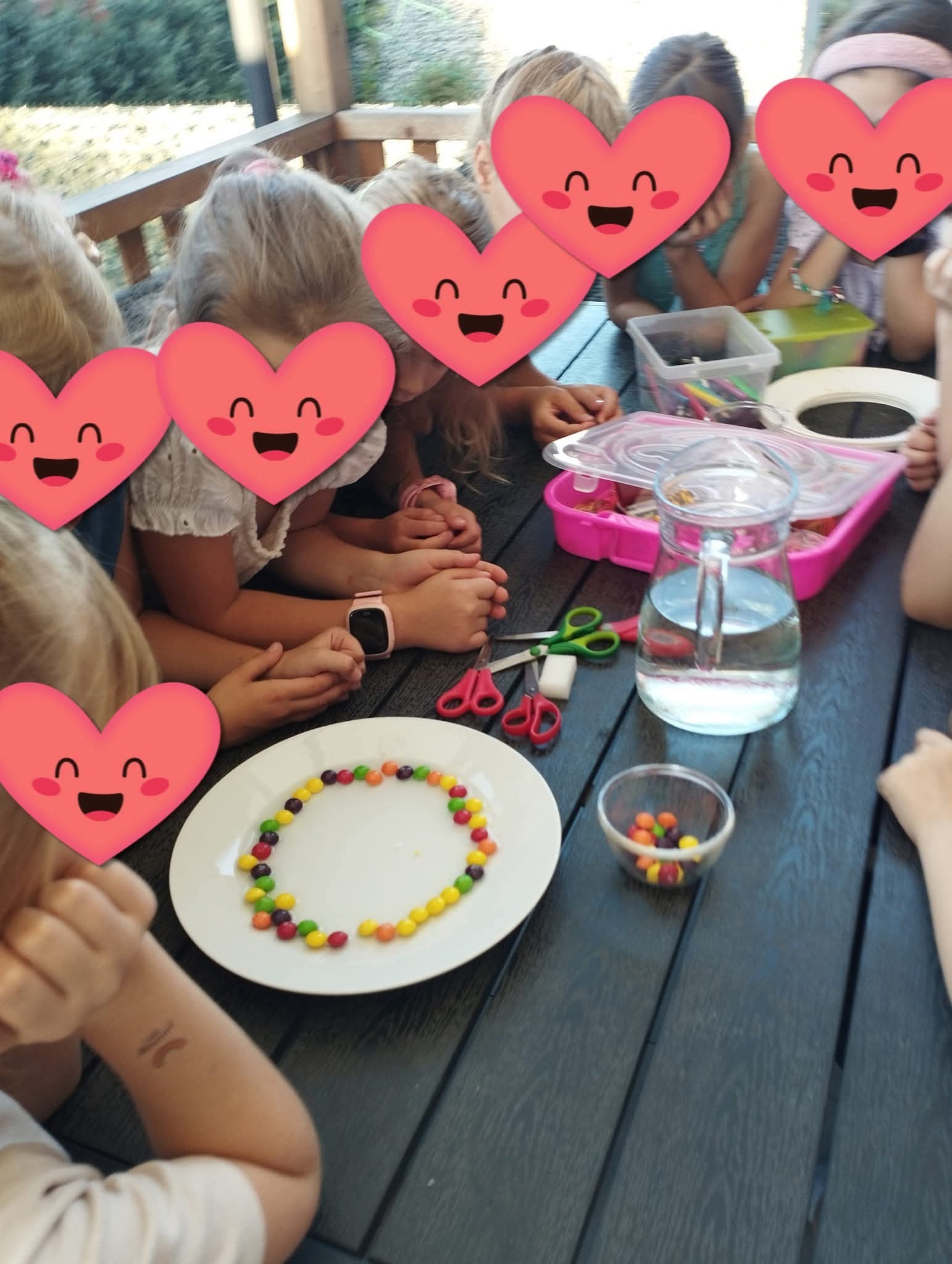 Dzieci przy stole układają kolorowe cukierki w kształt koła na białym talerzu. Widoczne nożyczki, dzbanek z wodą i pojemniki z przyborami.