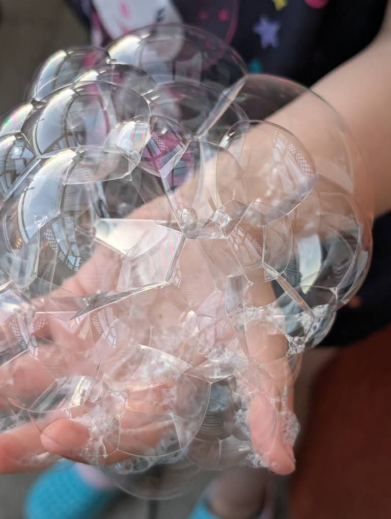 Dziecięca dłoń wypełniona kaskadą dużych, przezroczystych baniek mydlanych, tworzących geometryczny wzór. W tle fragment ubrania w gwiazdki i niebieskie buty.