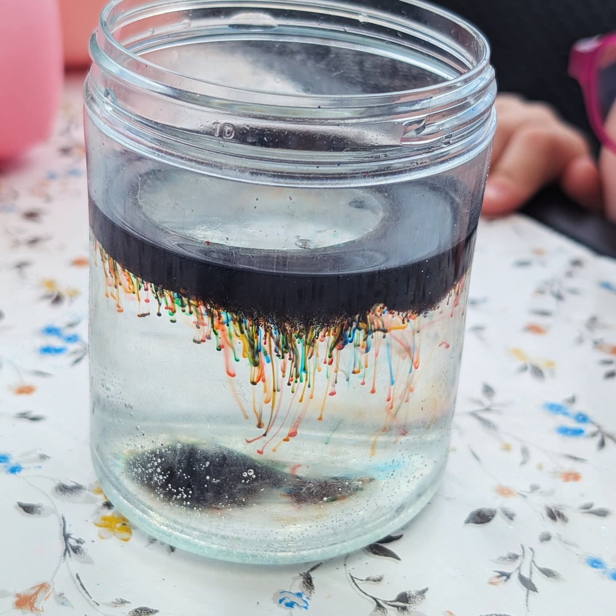 Eksperyment w słoiku: czarna ciecz z kolorowymi smugami opada w wodzie, tworząc efektowny wizualny pokaz. Widoczne bąbelki i wzorzyste tło.