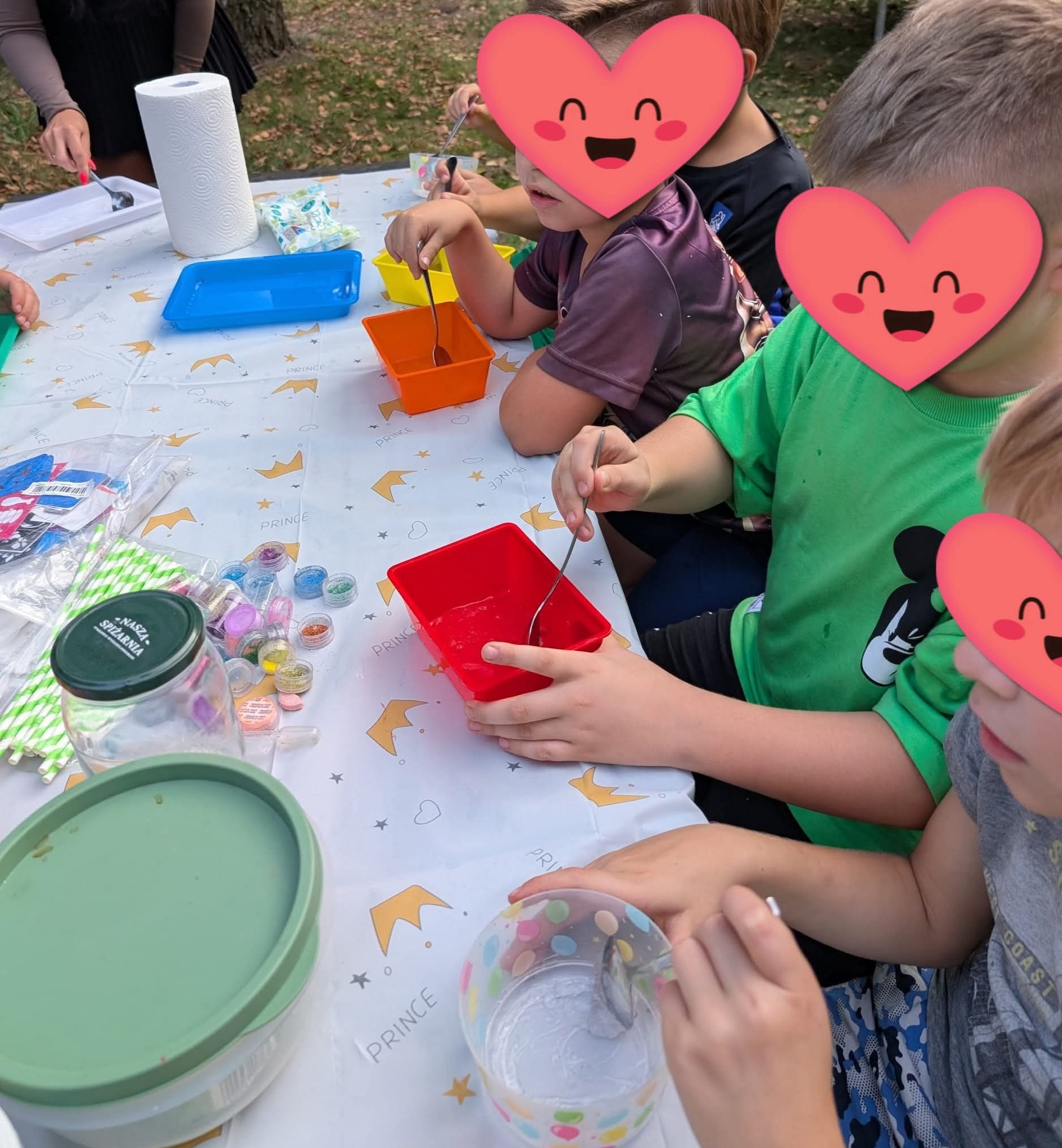 Warsztaty kreatywne dla dzieci: grupa bawi się przy stole, mieszając składniki w kolorowych pojemnikach. Widoczne brokaty, kleje i akcesoria do tworzenia slime.