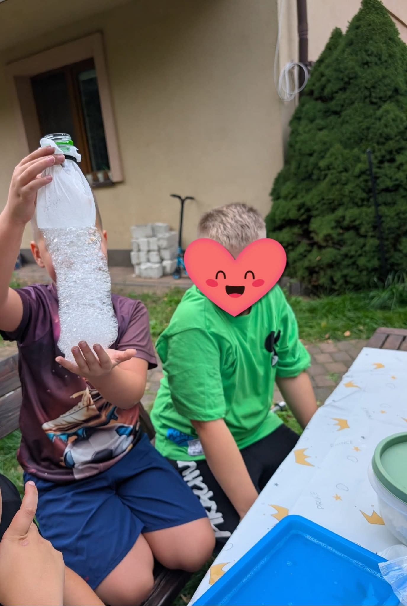 Chłopiec prezentuje butelkę wypełnioną pianą podczas zabawy na zewnątrz. Drugi chłopiec siedzi obok przy stole z dekoracyjnym obrusem. W tle zieleń ogrodu.