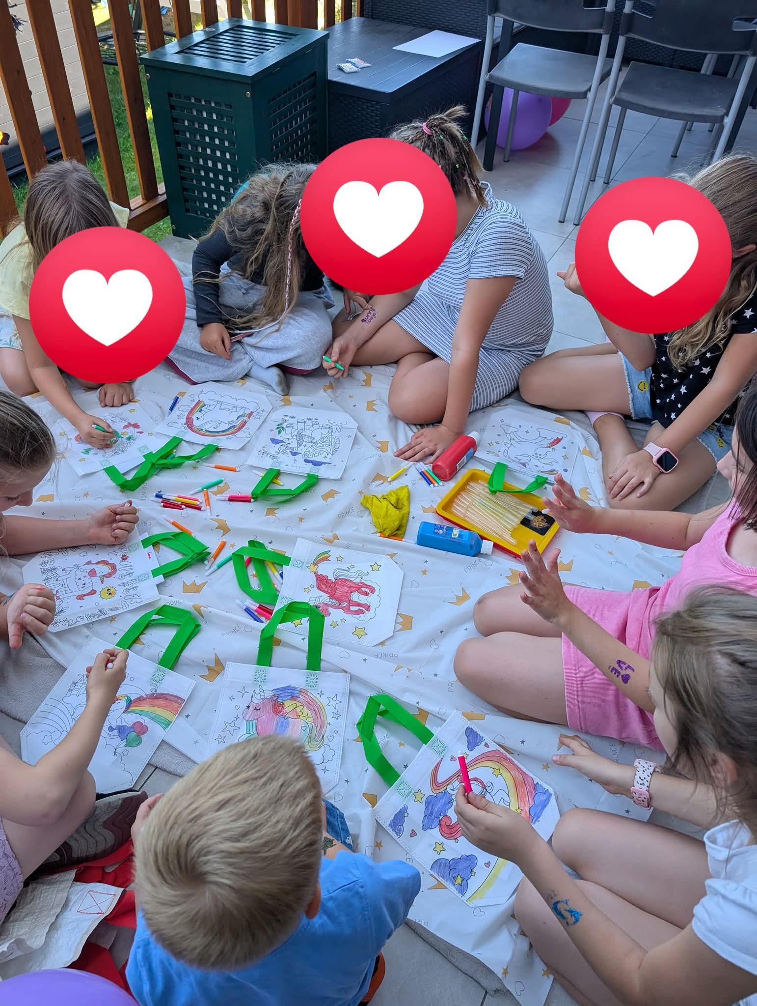 Dzieci siedzą na kocu, kolorując torby z jednorożcami podczas przyjęcia. Widoczne markery, farby i balony. Kreatywne zajęcie dla dzieci na imprezie.