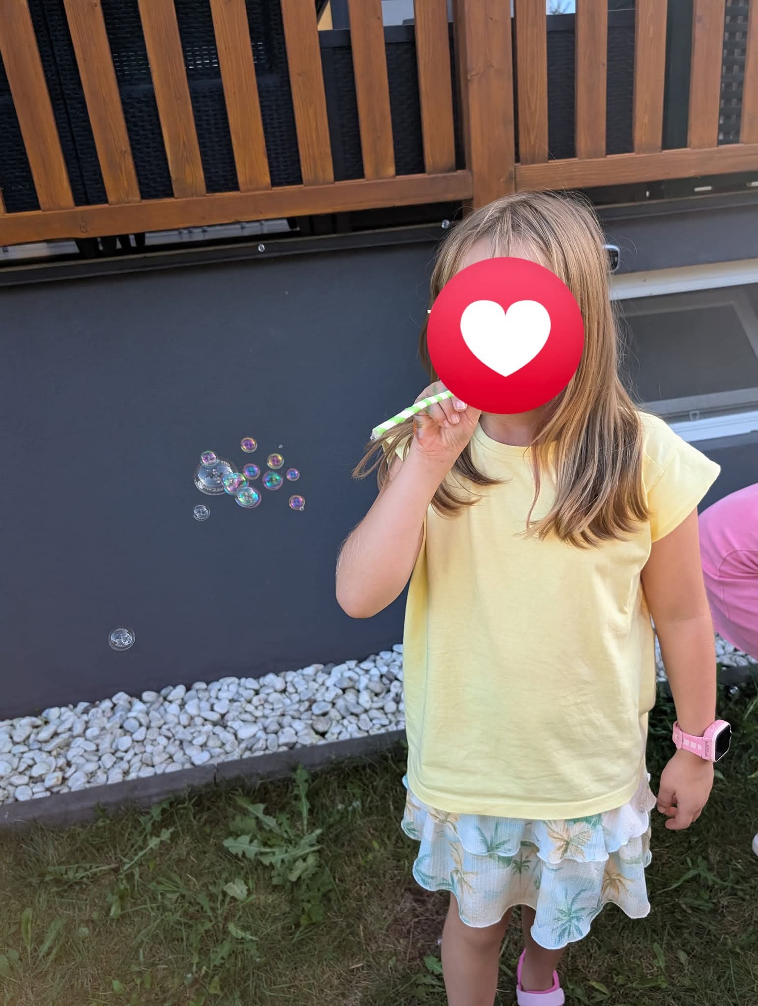 Dziewczynka w żółtej koszulce dmucha bańki mydlane na tle szarej ściany i kamieni. Widoczne tęczowe refleksy na bańkach. Ujęcie z zewnątrz, w dziennym świetle.