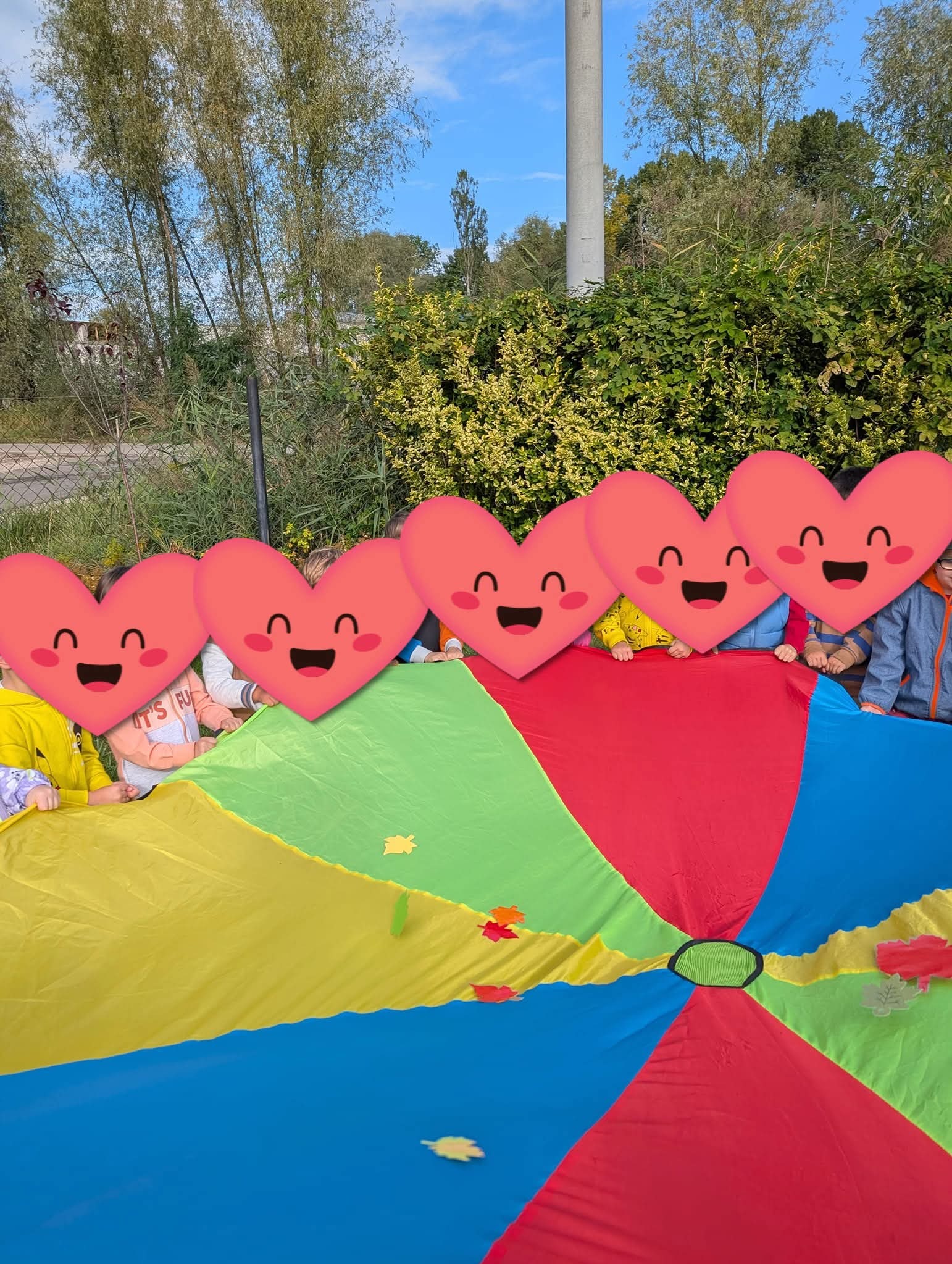 Grupka dzieci z zakrytymi twarzami w serduszka bawi się kolorową chustą animacyjną na tle zieleni. Jesienne liście na chuście. Zabawa na świeżym powietrzu.