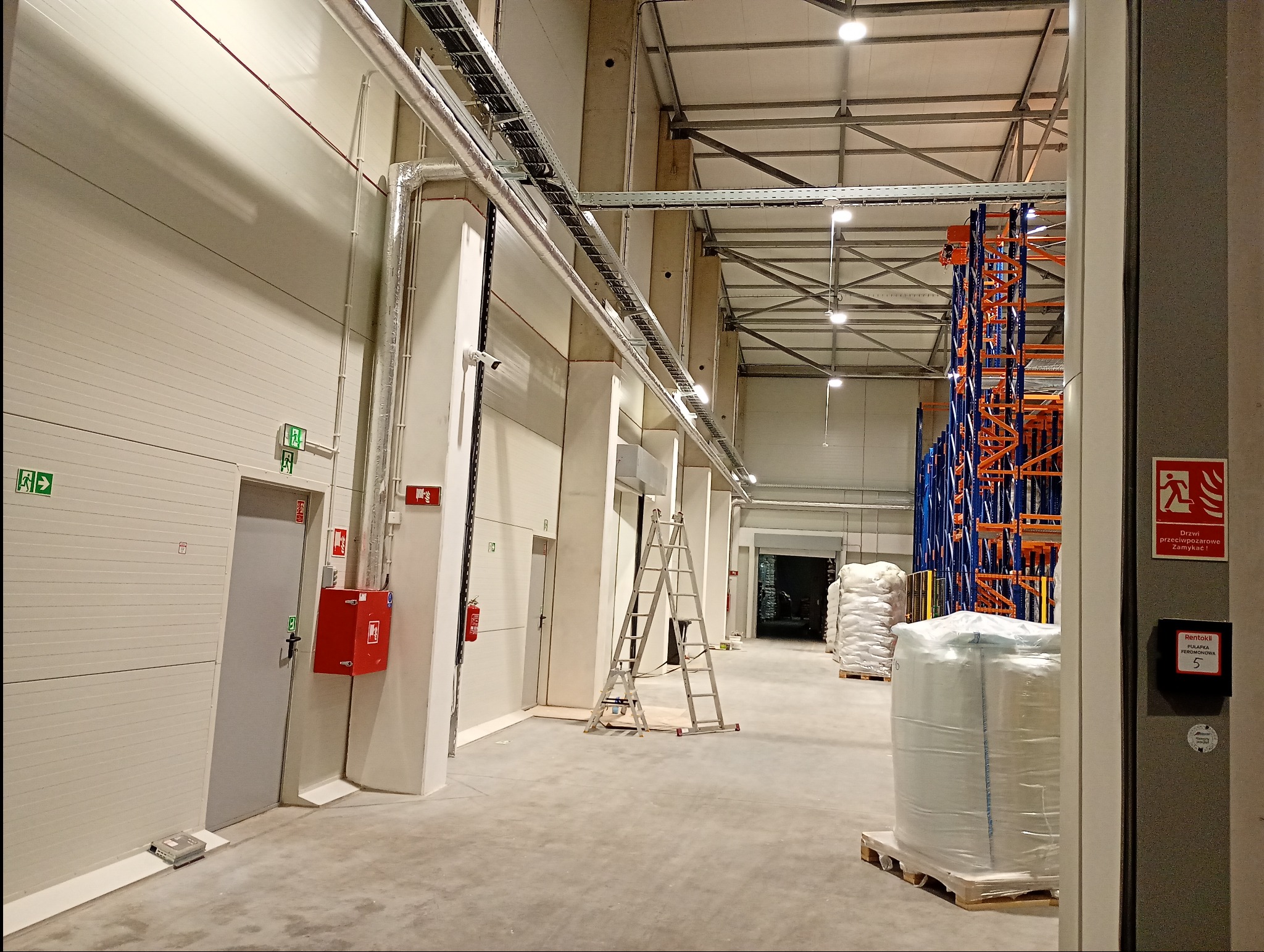 Wnętrze hali magazynowej po remoncie: białe ściany, instalacje, drabina, regały. Widoczne elementy systemu przeciwpożarowego i wentylacji. Czysta posadzka.