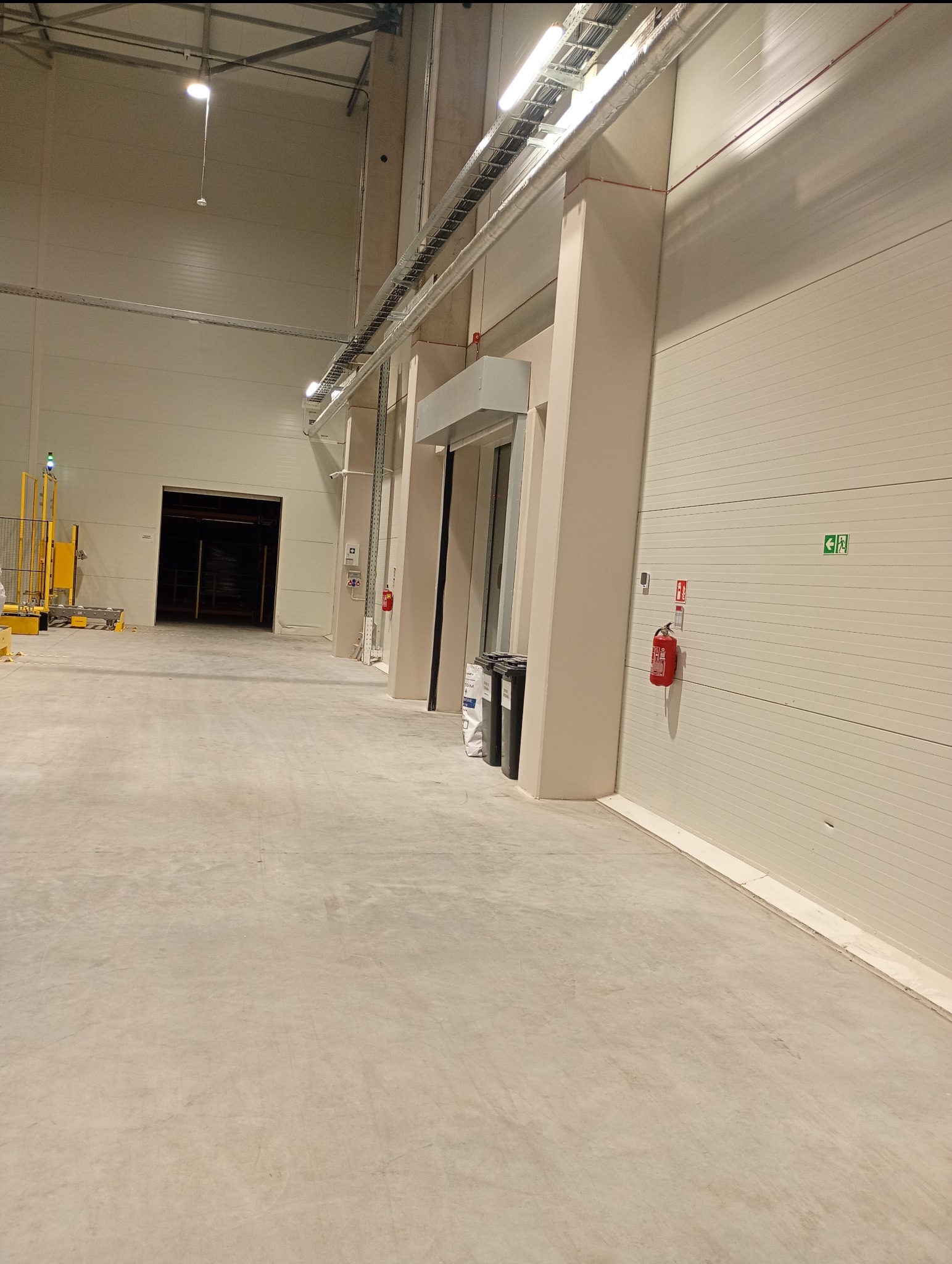 Wnętrze hali magazynowej z betonową posadzką i segmentową bramą. Widoczne instalacje pod sufitem, oświetlenie i sprzęt. Ściany w jasnym odcieniu beżu.