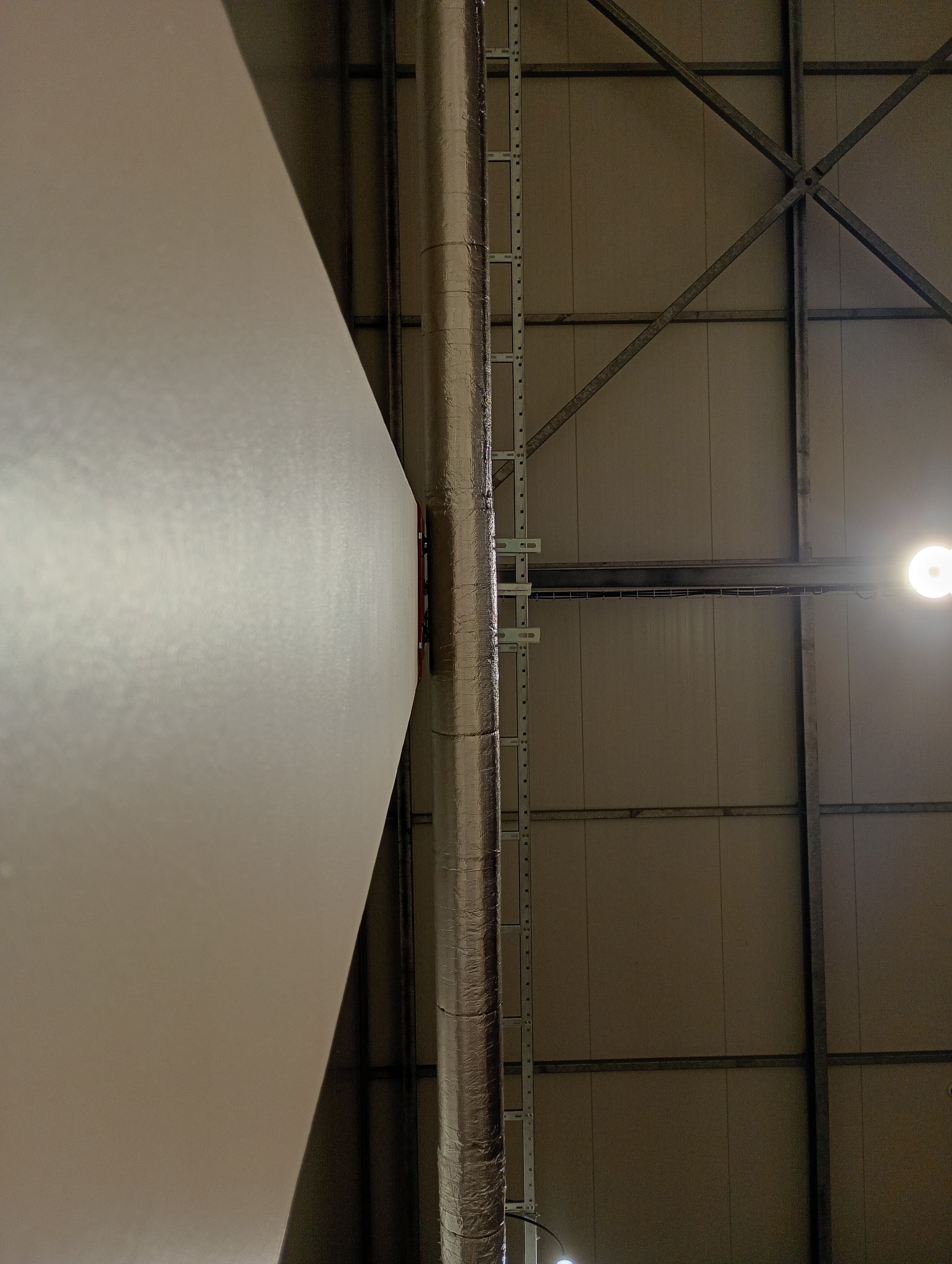 Sufit w hali produkcyjnej z widoczną płytą kartonowo-gipsową, rurą wentylacyjną w izolacji i elementami konstrukcji stalowej. Ujęcie z dołu, oświetlenie punktowe.