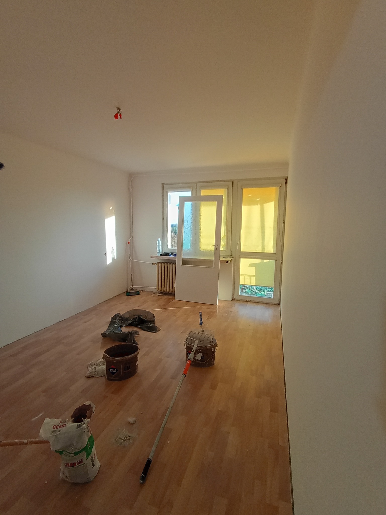 Pokój w trakcie remontu: widoczne narzędzia malarskie, wiadro z farbą, worki z gładzią, parkiet i przygotowane ściany. Dziennie światło wpada przez okno balkonowe.