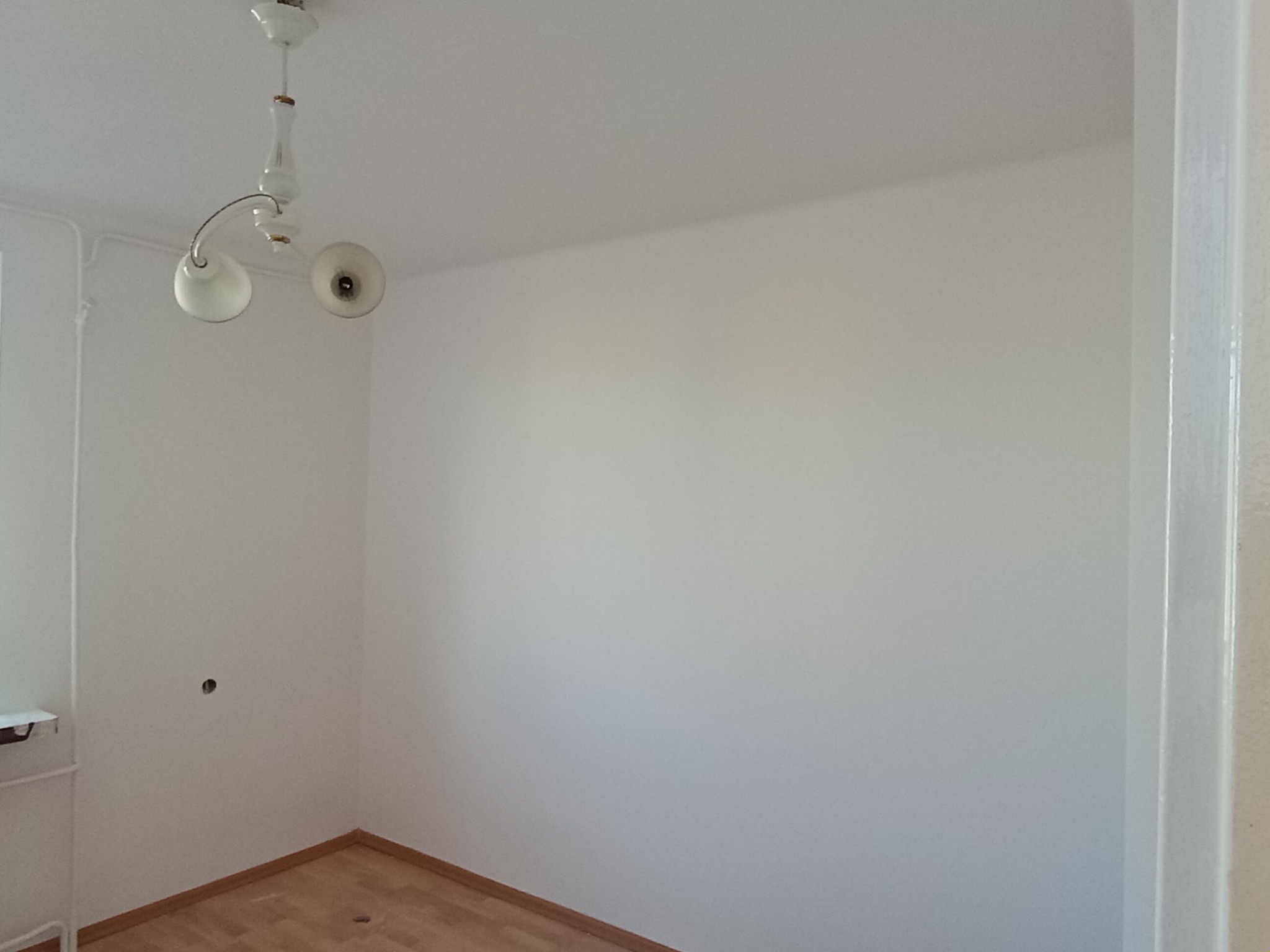 Pomalowane na biało wnętrze pokoju z drewnianą podłogą i lampą sufitową. Widoczna ściana z otworem na gniazdko i fragment białej futryny drzwi.