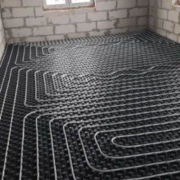 Instalacja ogrzewania podłogowego w surowym wnętrzu w Warszawie. Białe rury ułożone na czarnej macie, widoczne okno i ściany z bloczków.