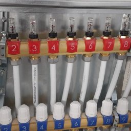 ARCHITECTURE OF TECHNOLOGY SPÓŁKA Z OGRANICZONĄ ODPOWIEDZIALNOŚCIĄ - Rozdzielacz ogrzewania podłogowego z dziewięcioma obwodami, z numeracją od 1 do 9, zamontowany w metalowej szafce. Widoczne białe rury i elementy regulacyjne.