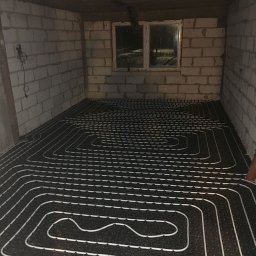 ARCHITECTURE OF TECHNOLOGY SPÓŁKA Z OGRANICZONĄ ODPOWIEDZIALNOŚCIĄ - Ułożona instalacja ogrzewania podłogowego w surowym pomieszczeniu z białej cegły. Widoczne okno i wystające przewody elektryczne. Czarna mata z białymi rurkami.