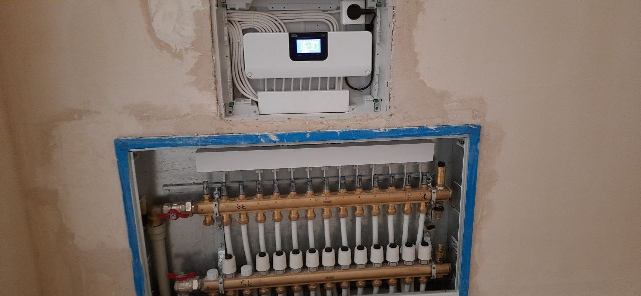 Rozdzielacz ogrzewania podłogowego z zaworami i biała skrzynka sterująca na ścianie w trakcie montażu, widoczne przewody i rury. Ściana w stanie surowym.