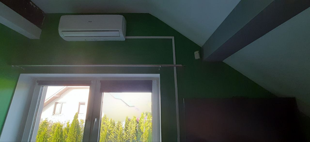 Klimatyzator na zielonej ścianie pod skosem, widoczne rury instalacyjne i okno z widokiem na zieleń. Ujęcie z dołu, podkreślające montaż w nietypowym miejscu.