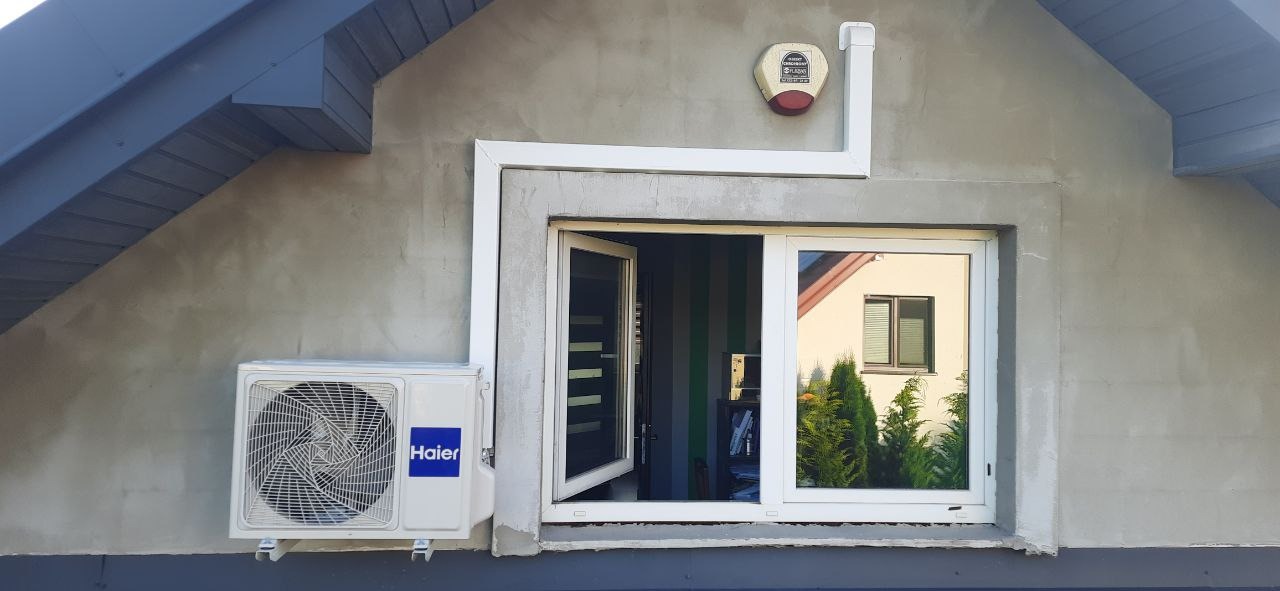 Biały klimatyzator Haier zamontowany na ścianie budynku obok okna. Widoczny alarm na elewacji i fragment dachu. Ściana w surowym tynku.