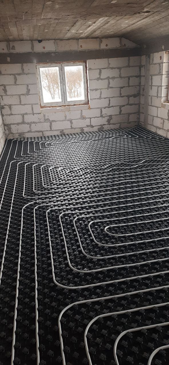 Instalacja ogrzewania podłogowego w surowym wnętrzu w Warszawie. Białe rury ułożone na czarnej macie, widoczne okno i ściany z bloczków.