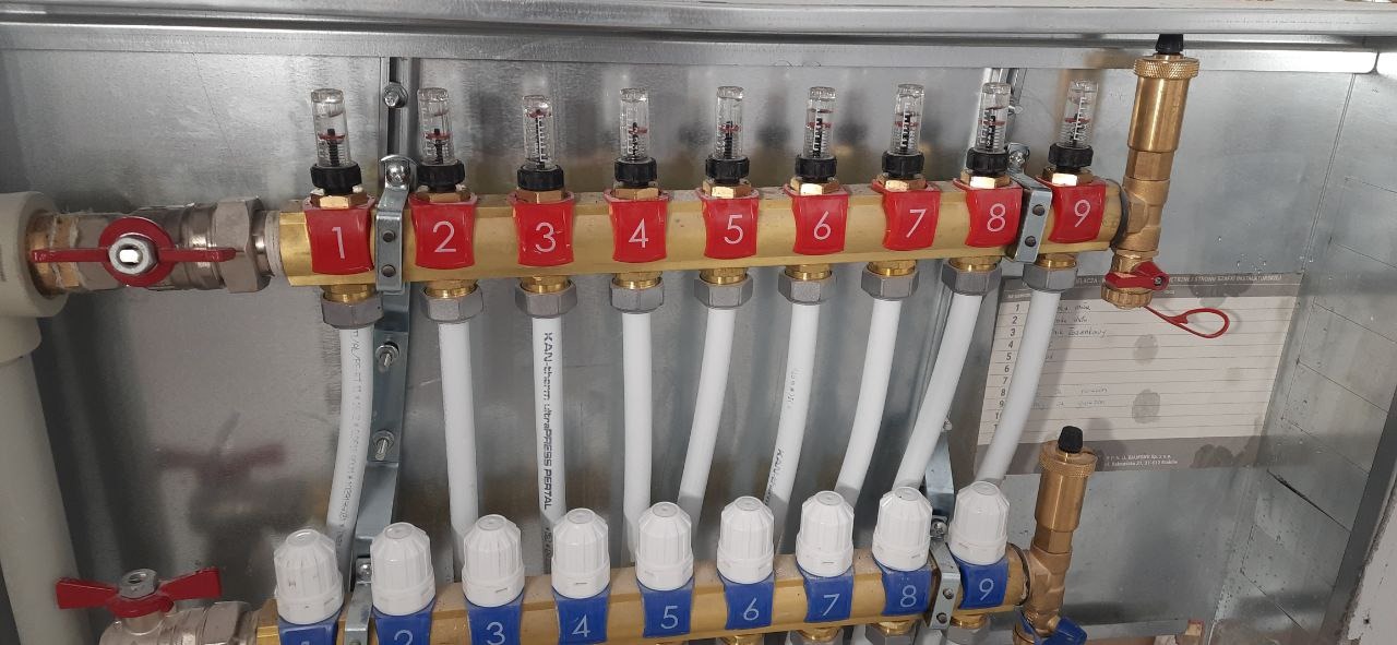 Rozdzielacz ogrzewania podłogowego z dziewięcioma obwodami, z numeracją od 1 do 9, zamontowany w metalowej szafce. Widoczne białe rury i elementy regulacyjne.