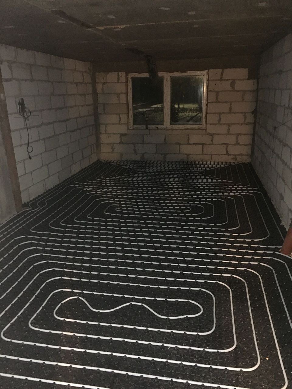 Ułożona instalacja ogrzewania podłogowego w surowym pomieszczeniu z białej cegły. Widoczne okno i wystające przewody elektryczne. Czarna mata z białymi rurkami.
