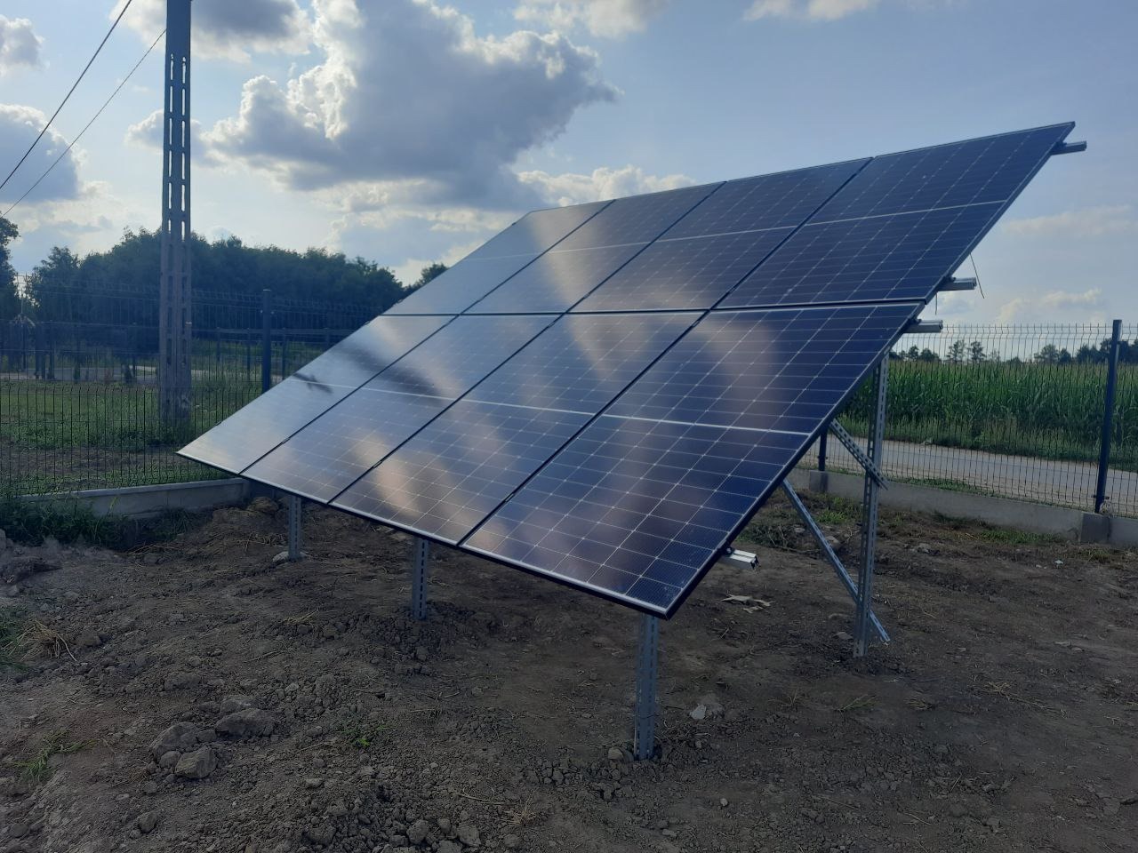 Ustawiona pod kątem instalacja fotowoltaiczna na metalowej konstrukcji, widoczna na tle błękitnego nieba z chmurami i zieleni. Panele odbijają niebo.