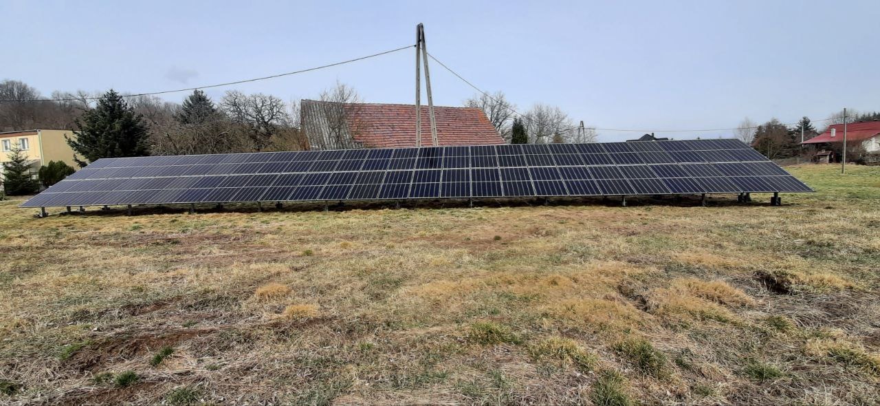 Długa instalacja fotowoltaiczna na łące, widok z daleka. Panele ustawione poziomo, w tle budynki mieszkalne i słup energetyczny. Realizacja w wiejskim otoczeniu.