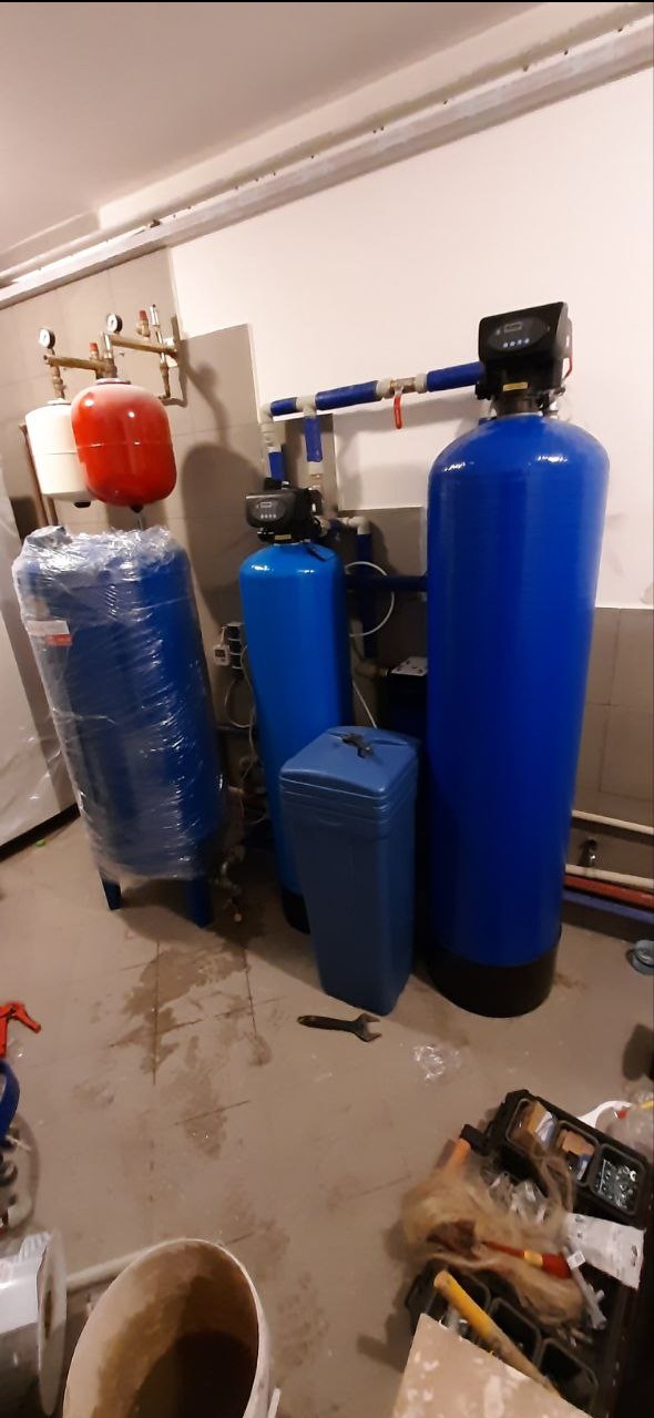Niebieskie filtry wody w piwnicy, jeden owinięty folią. Manometry na ścianie, narzędzia na podłodze. Instalacja wodna w trakcie prac.