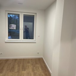 Andrii Tovstyk - Jasne, puste wnętrze po remoncie: drewniana podłoga, białe ściany, okno z odbiciem osoby. Minimalistyczny design, świeżo wykończone.