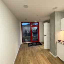 Andrii Tovstyk - Jasne wnętrze po remoncie: świeżo pomalowane ściany, drewniana podłoga, białe drzwi i sufit z wbudowanymi głośnikami. Widoczne wejście z czerwoną ramą.