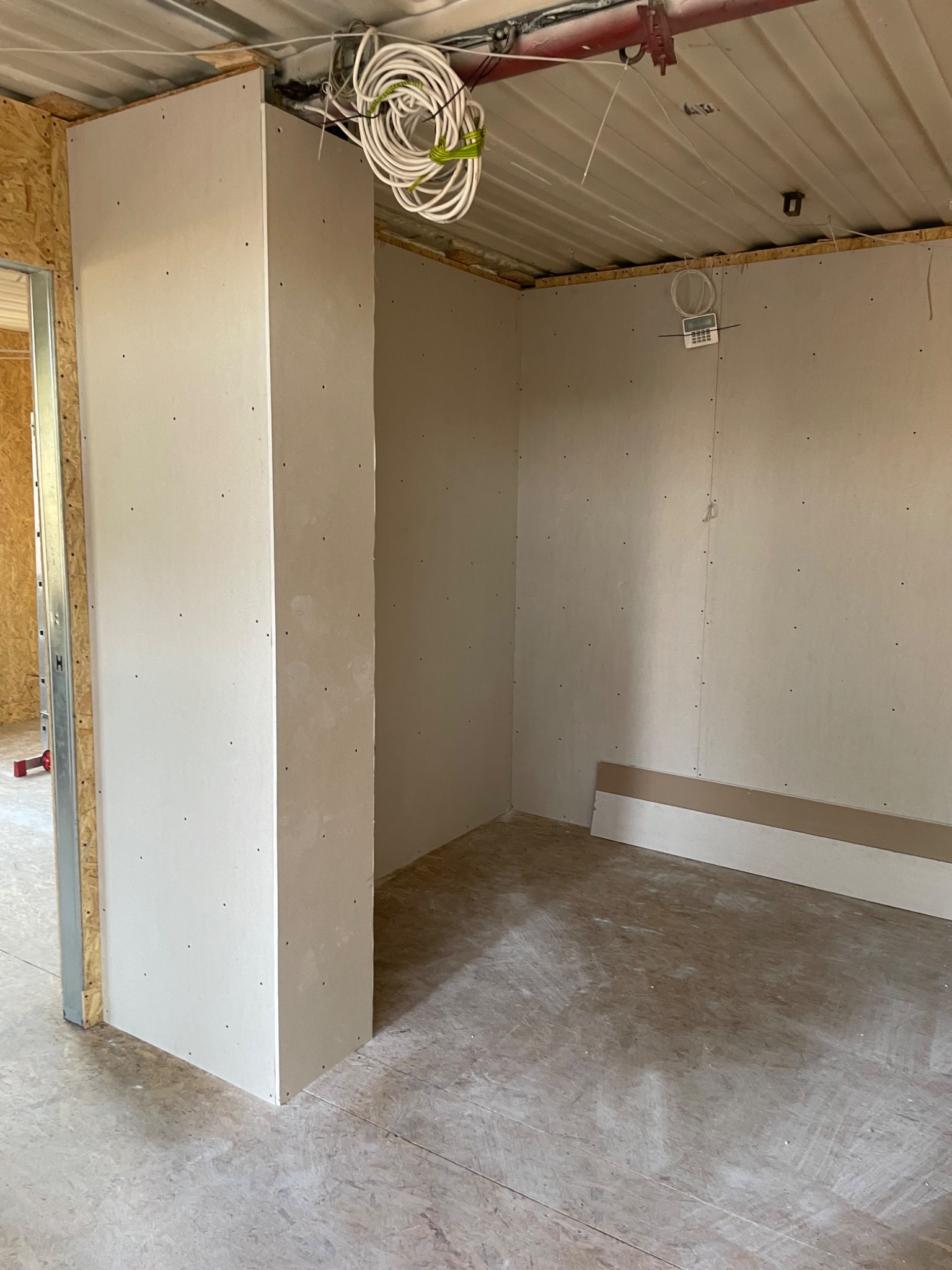 Wnętrze w trakcie prac wykończeniowych: ściany z płyt karton-gips, widoczne przewody elektryczne pod sufitem, surowa podłoga. Fragment metalowej konstrukcji drzwi.