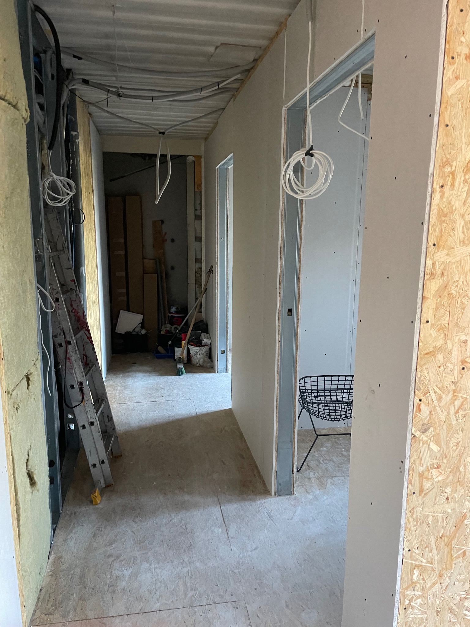Korytarz w trakcie prac wykończeniowych: płyty karton-gips na ścianach, widoczne instalacje elektryczne, drabina i narzędzia. Surowy stan, przygotowanie do dalszych prac.