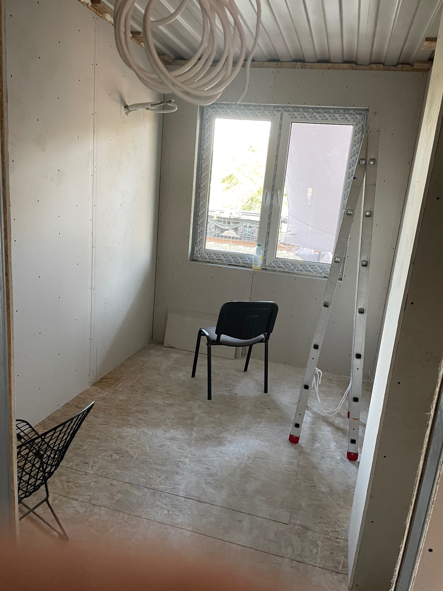 Pokój w trakcie wykańczania: ściany z płyt karton-gips, okno z widokiem na zewnątrz, drabina oparta o ścianę, krzesło. Widoczne przewody elektryczne pod sufitem.