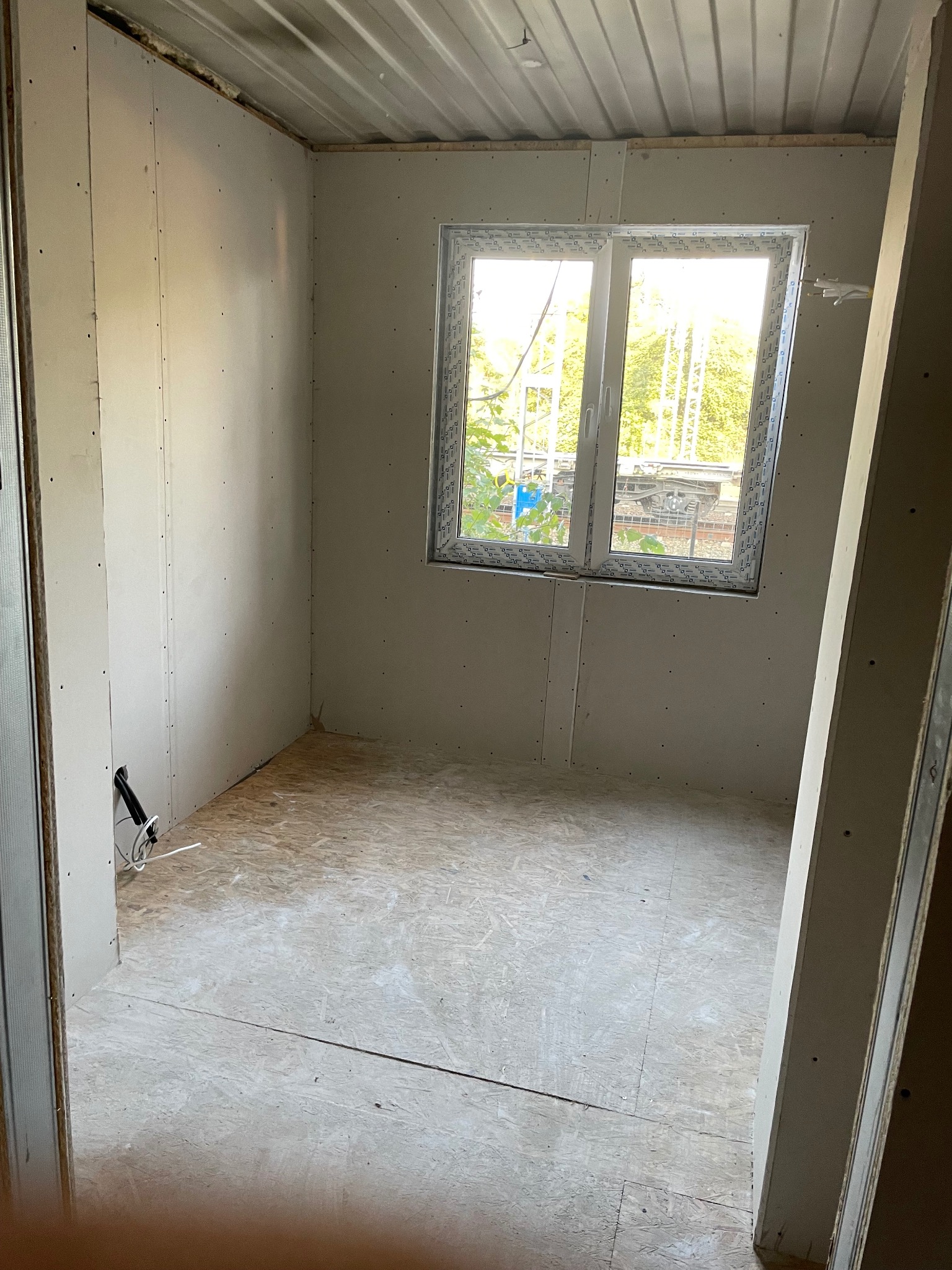 Pokój w trakcie wykańczania: ściany z płyt kartonowo-gipsowych, okno z widokiem na zieleń, podłoga z płyt OSB. Widoczne przewody elektryczne i fragment ościeżnicy.