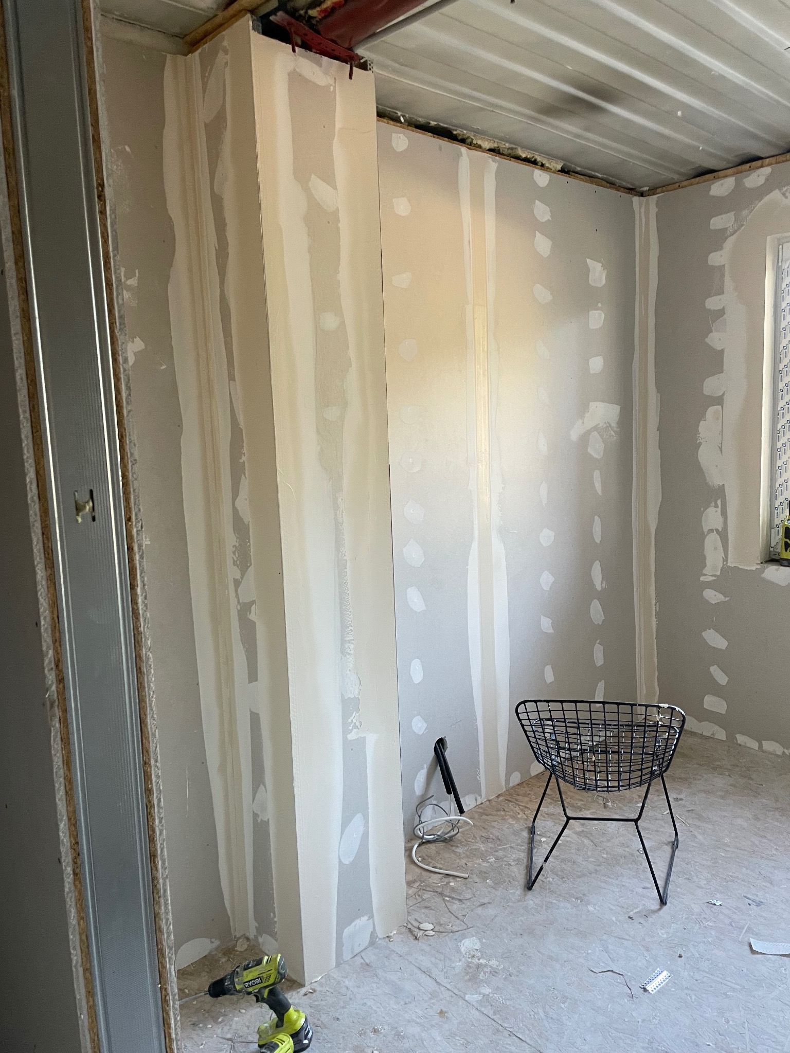 Wnętrze w trakcie prac wykończeniowych: ściany z płyt karton-gips, widoczne szpachlowanie, metalowe profile, narzędzia i krzesło. Surowy stan, przygotowanie do dalszych prac.