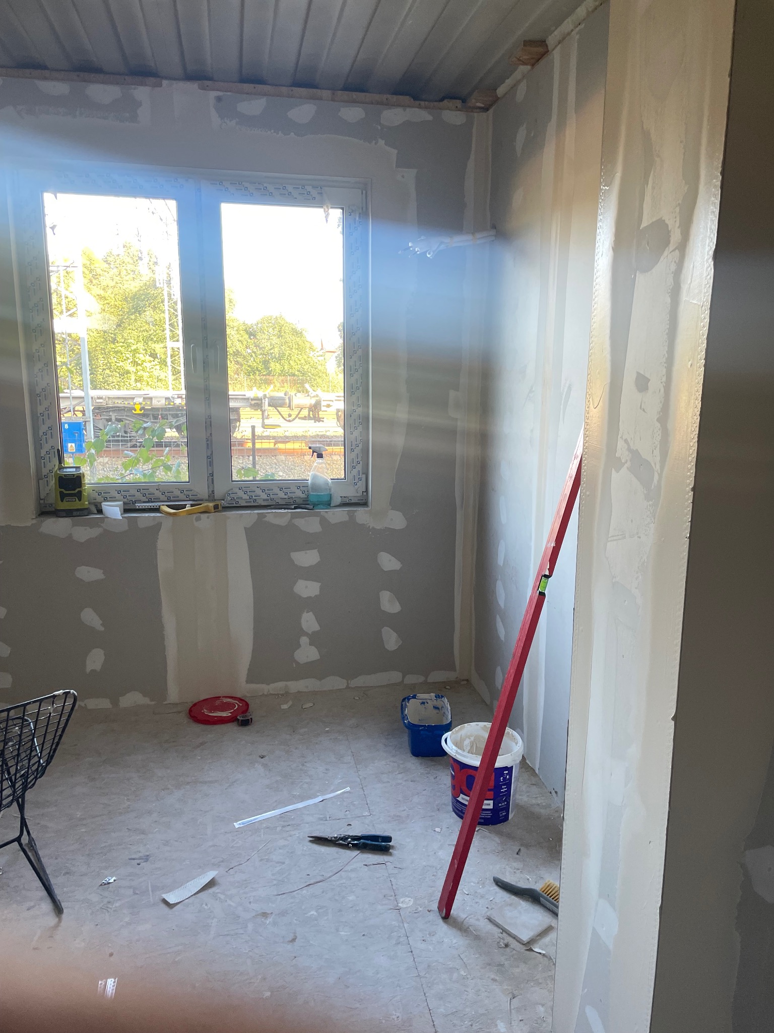 Wnętrze w trakcie szpachlowania: ściany z płyt gipsowo-kartonowych z widocznymi punktami szpachli, okno z widokiem na zewnątrz, narzędzia i poziomica. Prace wykończeniowe w toku.