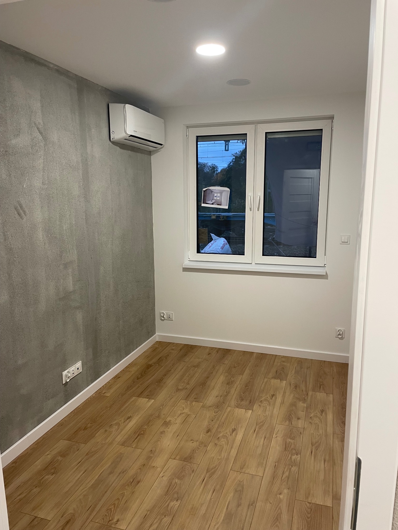 Pokój po remoncie: szara ściana z klimatyzatorem, drewniana podłoga, białe okno z widokiem na zewnątrz. Minimalistyczny design, świeżo wykończone wnętrze.