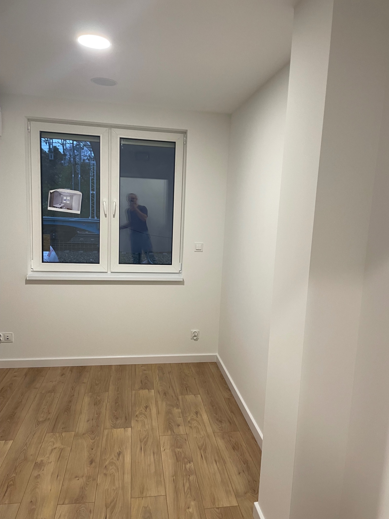 Jasne, puste wnętrze po remoncie: drewniana podłoga, białe ściany, okno z odbiciem osoby. Minimalistyczny design, świeżo wykończone.