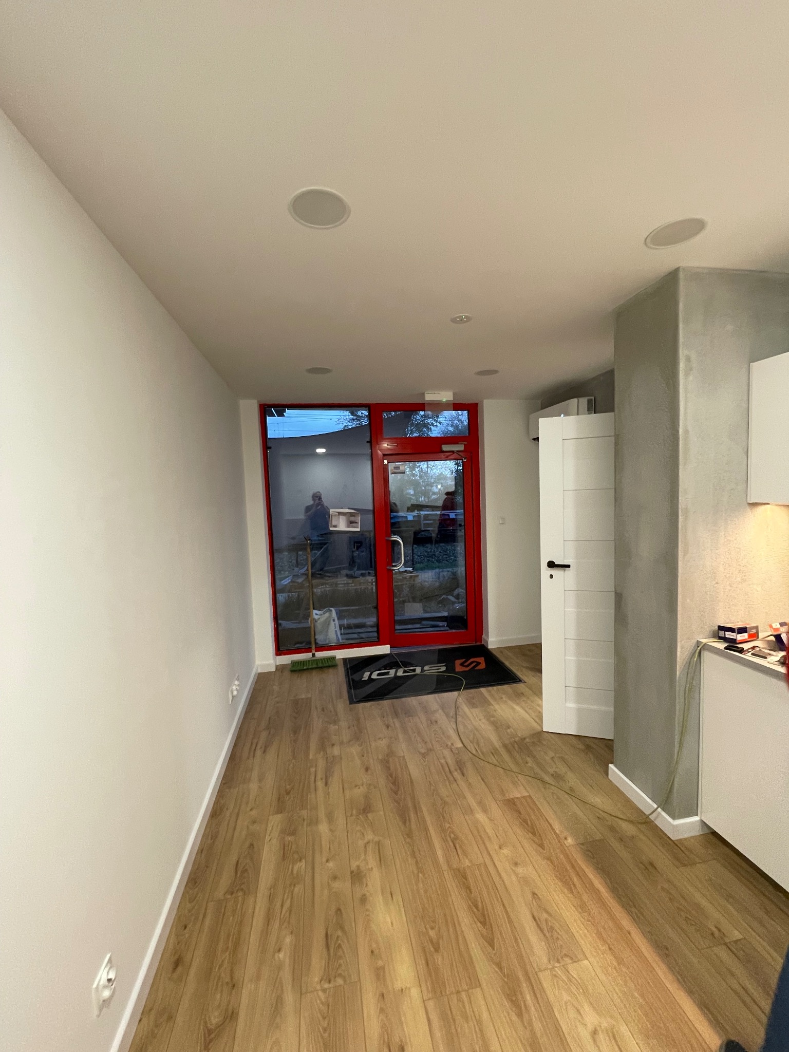 Jasne wnętrze po remoncie: świeżo pomalowane ściany, drewniana podłoga, białe drzwi i sufit z wbudowanymi głośnikami. Widoczne wejście z czerwoną ramą.