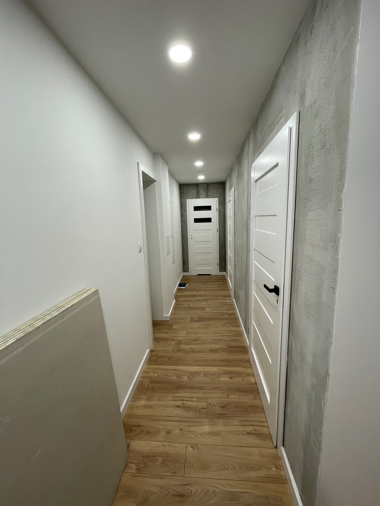 Wykończony korytarz z drewnopodobną podłogą, białymi drzwiami i szarą ścianą o fakturze betonu. Widoczne płyty gipsowe przy ścianie.