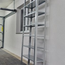 Tomasz Wójcik - Metalowy regał z półkami, ustawiony przy białej ścianie w garażu. Widoczne okno i brama garażowa. Podłoga wyłożona kostką brukową.