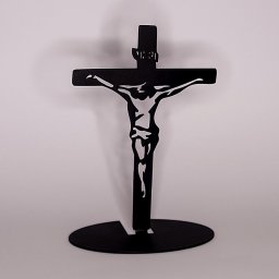 Tomasz Wójcik - Czarny, metalowy krucyfiks z wyciętą sylwetką Jezusa na owalnej podstawie, stojący na jasnym tle. Precyzyjne cięcie, minimalistyczny design.