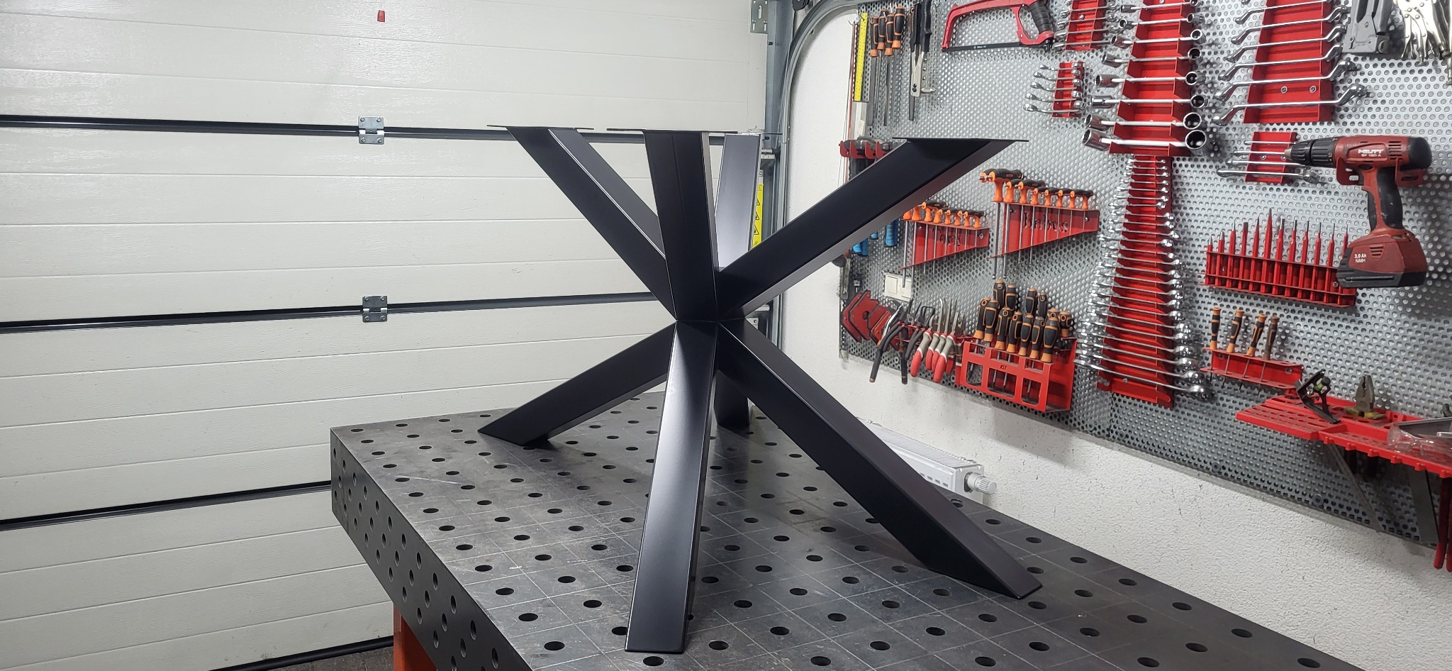 Czarny, metalowy stelaż stołu o geometrycznym kształcie na perforowanym stole warsztatowym. W tle ściana z narzędziami i brama garażowa.
