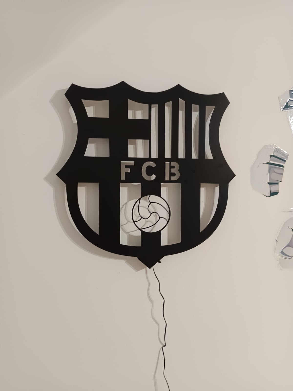 Czarny, ażurowy herb FCB z piłką, wykonany z metalu, podświetlony, zawieszony na białej ścianie. Precyzyjne cięcie, nowoczesny design.
