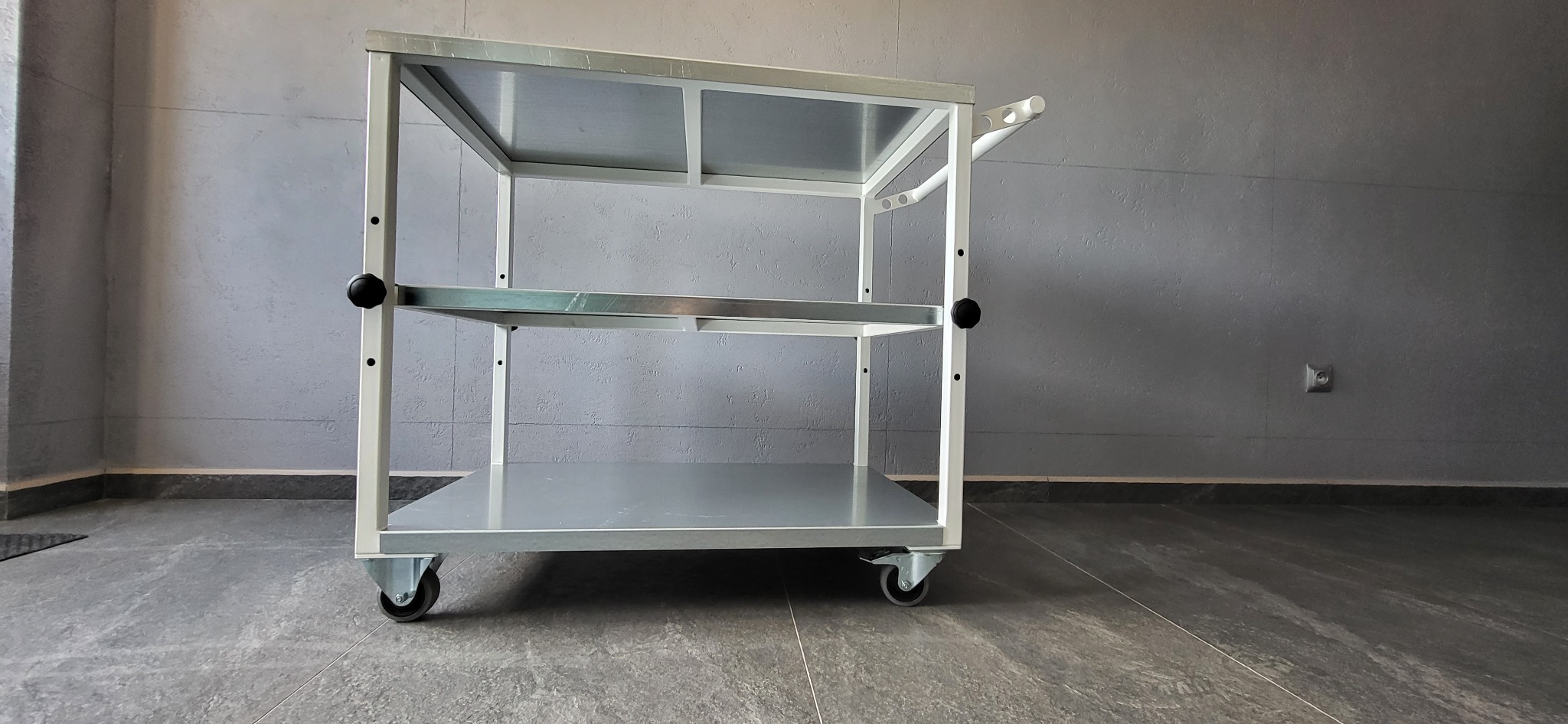 Metalowy wózek warsztatowy na kółkach, z trzema półkami, spawany, z uchwytem do prowadzenia. Widoczne ślady obróbki. Tło stanowi szara ściana i podłoga.