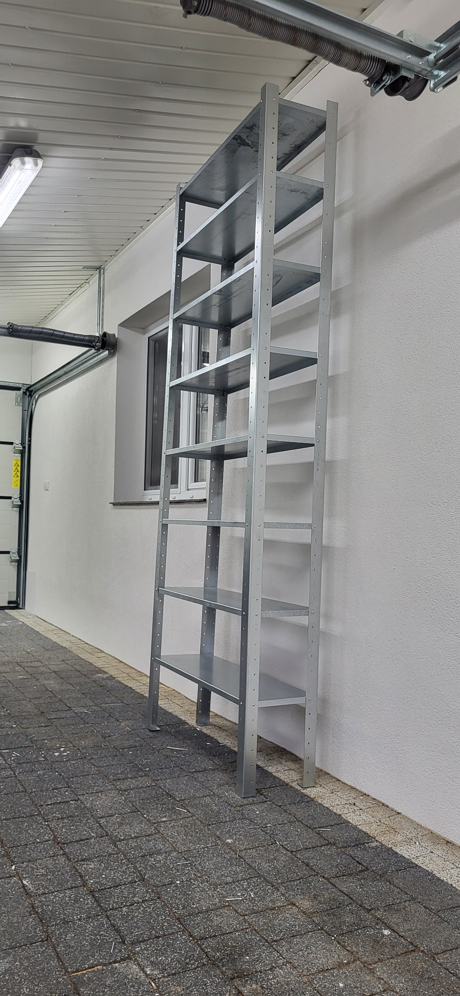 Metalowy regał z półkami, ustawiony przy białej ścianie w garażu. Widoczne okno i brama garażowa. Podłoga wyłożona kostką brukową.