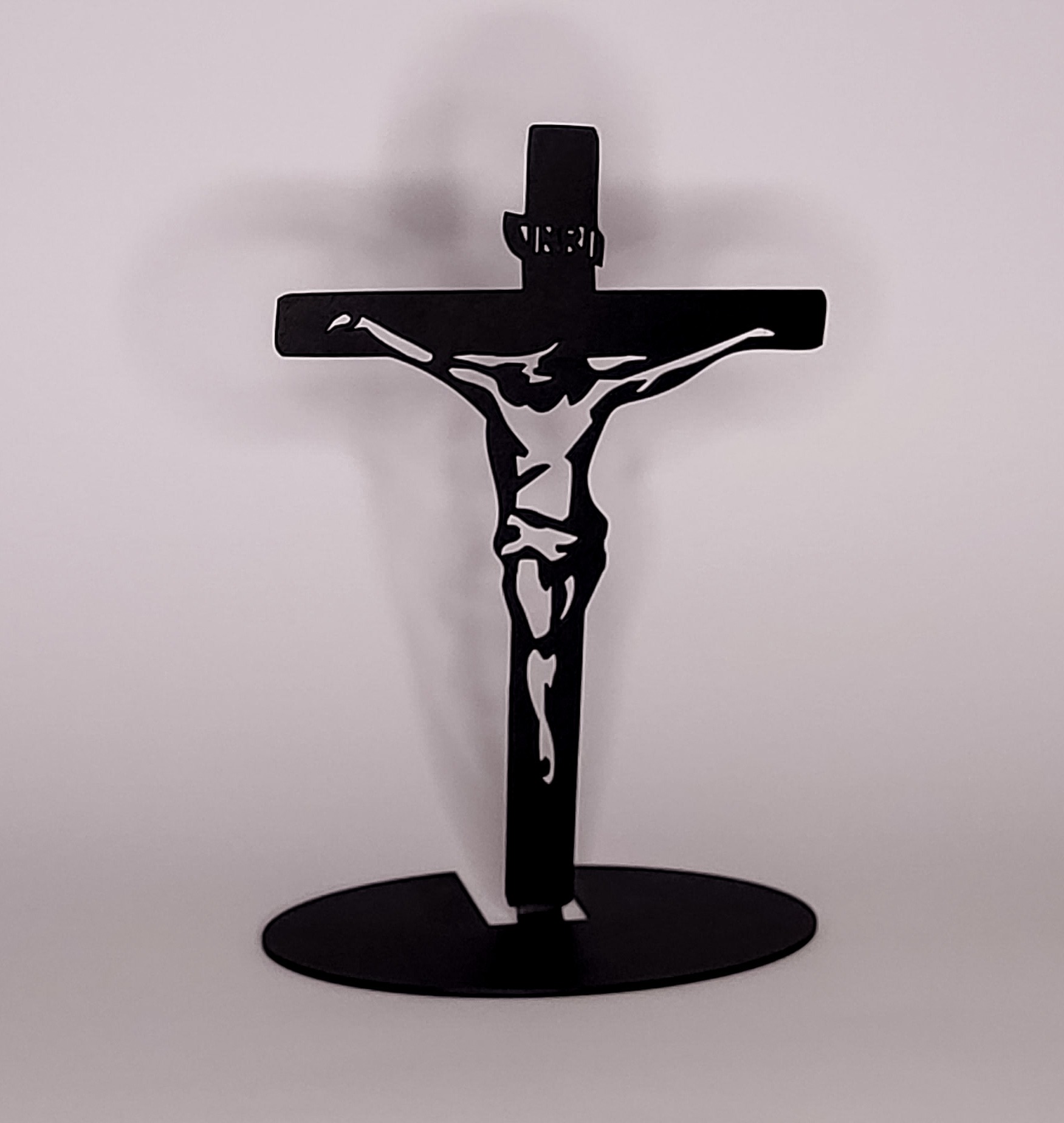 Czarny, metalowy krucyfiks z wyciętą sylwetką Jezusa na owalnej podstawie, stojący na jasnym tle. Precyzyjne cięcie, minimalistyczny design.